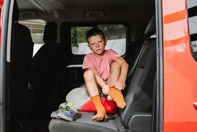 Full body kid in pink t shirt putting on orange socks and looking at camera while sitting on back seat of car during road trip