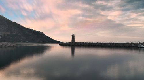 Lighthouse by river and buildings against sky during sunset