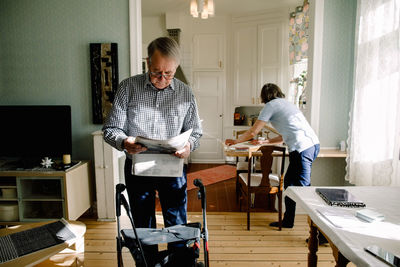 Retired senior man reading newspaper while female caregiver working in kitchen at nursing home