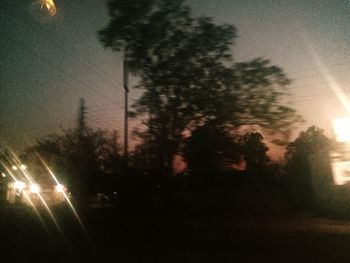 Silhouette trees by illuminated street against sky at dusk