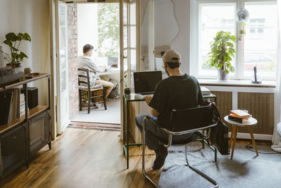 Rear view of gay couple using laptops while sitting on chairs at home