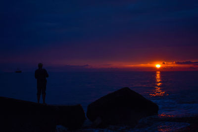 Rear view of silhouette man standing on rock by sea against sky during sunset
