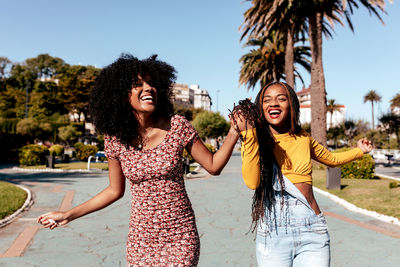 Delighted black woman with braids holding hand of african american female best friend with curly hair while walking along street in tropical city on sunny day