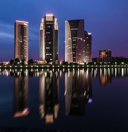 Illuminated modern buildings by river against sky in city at dusk