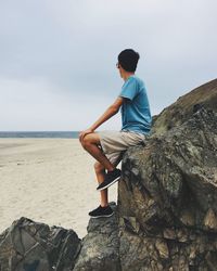 Side view full length of young man sitting on rock at beach