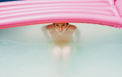 Boy in a pool taking a bath under a pink flat looking at camera.
