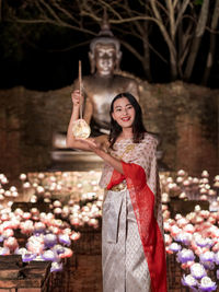 Portrait of smiling young woman with arms raised standing against lake
