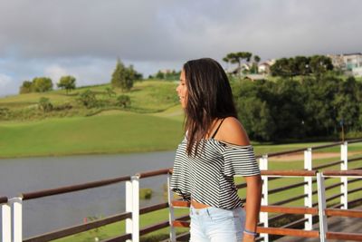 Young woman looking away while standing against river during sunny day