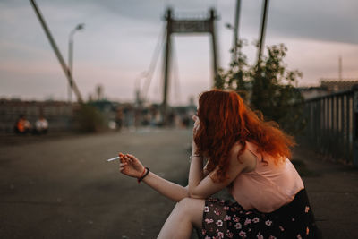 Young redheaded woman smoking cigarette while sitting on bridge