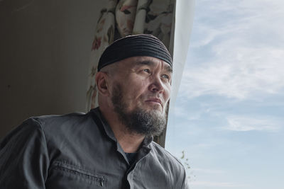 Senior asian muslim man with beard wearing skullcap sitting by window and looking into distance. 
