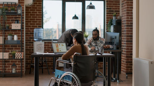 Rear view of woman sitting in wheelchair and using laptop at desk