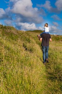 Father carrying daughter on shoulders while walking on grassy field against sky