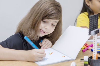 Close-up of girl drawing in book with friend while sitting in classroom at school