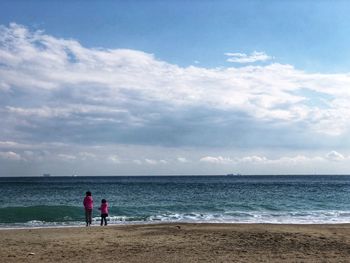 Rear view of sisters standing at beach against cloudy sky