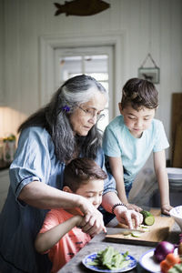 Grandmother assisting boy cutting cucumber on board in kitchen at home