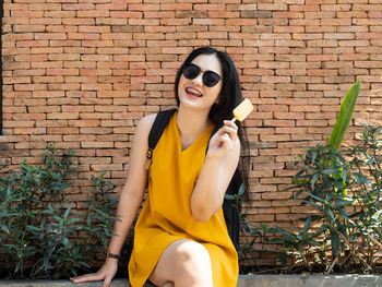 Young woman wearing sunglasses sitting against brick wall