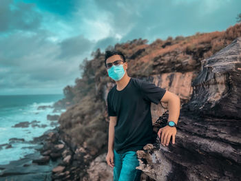 Young man wearing mask standing on rock by sea against sky