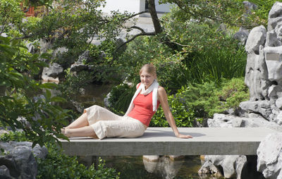 Young woman sitting on footbridge over lake amidst trees at garden