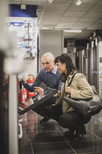 Mature owner explaining about oven to smiling female customer while crouching in electronics store