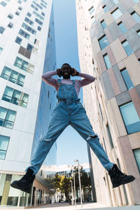 From below of young african american male in cool outfit jumping above ground on street while listening to songs in headphones and looking at camera