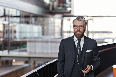 Serious mature businessman wearing headphones while talking through smart phone in office lobby