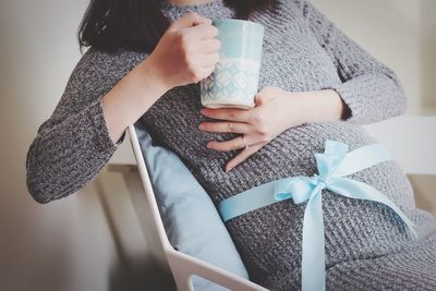 Midsection of pregnant woman sitting on chair with coffee at home