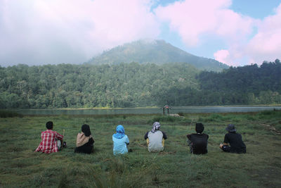 Rear view of people sitting on field by mountain against sky