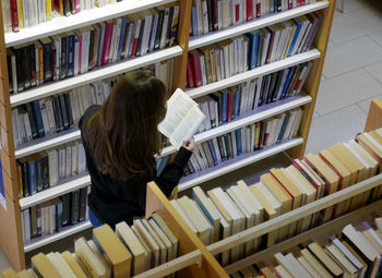 High angle view of woman reading book by shelf at library