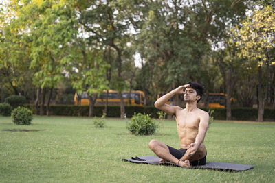 Full length of shirtless man sitting on grass against trees