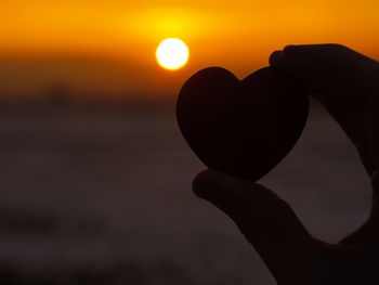 Cropped hand holding heart shape stone against sky during sunset