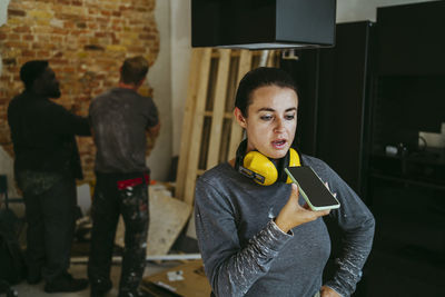 Female construction manager talking through smart phone speaker at site while workers working in background