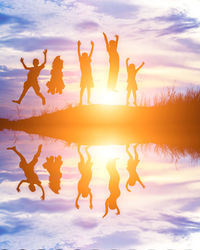 Silhouette children jumping at lakeshore against sky during sunset