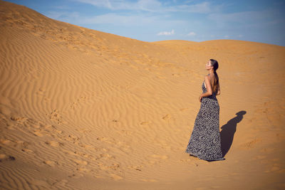 Brunette woman in a long leopard dress stands with her back in the desert at sunset. go everywhere