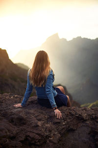 Woman sitting on rock against sky during sunset