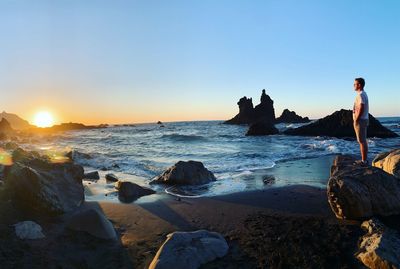 Side view of young man standing on rock formation at beach during sunset