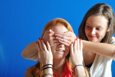 Young woman closing eyes of friend against blue background