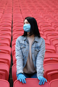 A female fan in a medical mask and rubber gloves sits alone in an empty stadium with red seats.