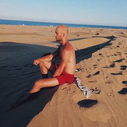 Full length of shirtless man sitting on sand at beach