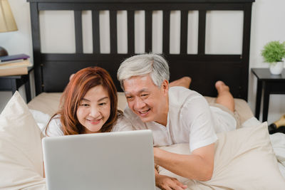 Smiling mature couple using laptop while lying on bed at home
