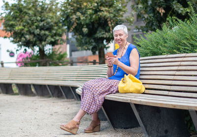 Aged woman sits on a bench in a public place in the city and uses a mobile phone