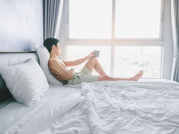 Asian young man with tattoo holding coffee cup on the bed looking out window in morning with sunrise 