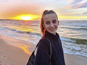 Portrait of young woman standing at beach against sky during sunset