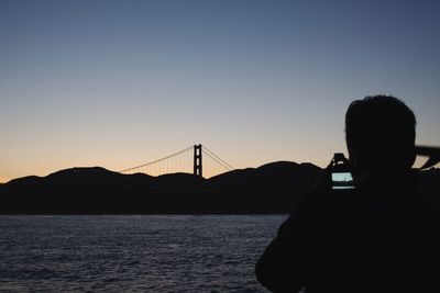 Silhouette tourist photographing golden gate bridge through smart phone against clear sky during sunset
