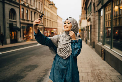 Happy young muslim woman taking selfie standing on sidewalk in city