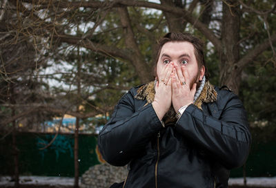 Shocked man with hands covering mouth against trees during winter at park