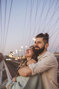 Boyfriend embracing happy girlfriend while standing on bridge in city