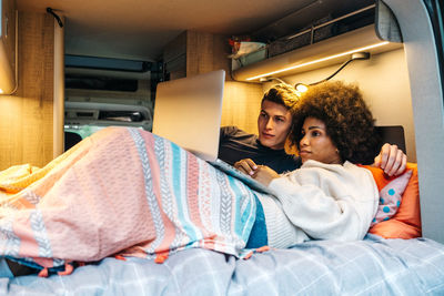 Happy romantic multiracial couple cuddling and enjoying time together while lying on bed and watching movie on laptop during travel in camper van