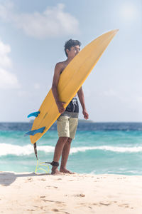 Rear view of shirtless young man with surfboard standing at beach against sky