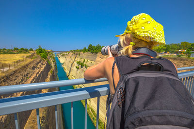 Rear view of woman photographing while standing on bridge against clear blue sky