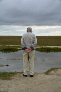 Rear view of senior man standing on field looking at pond against sky